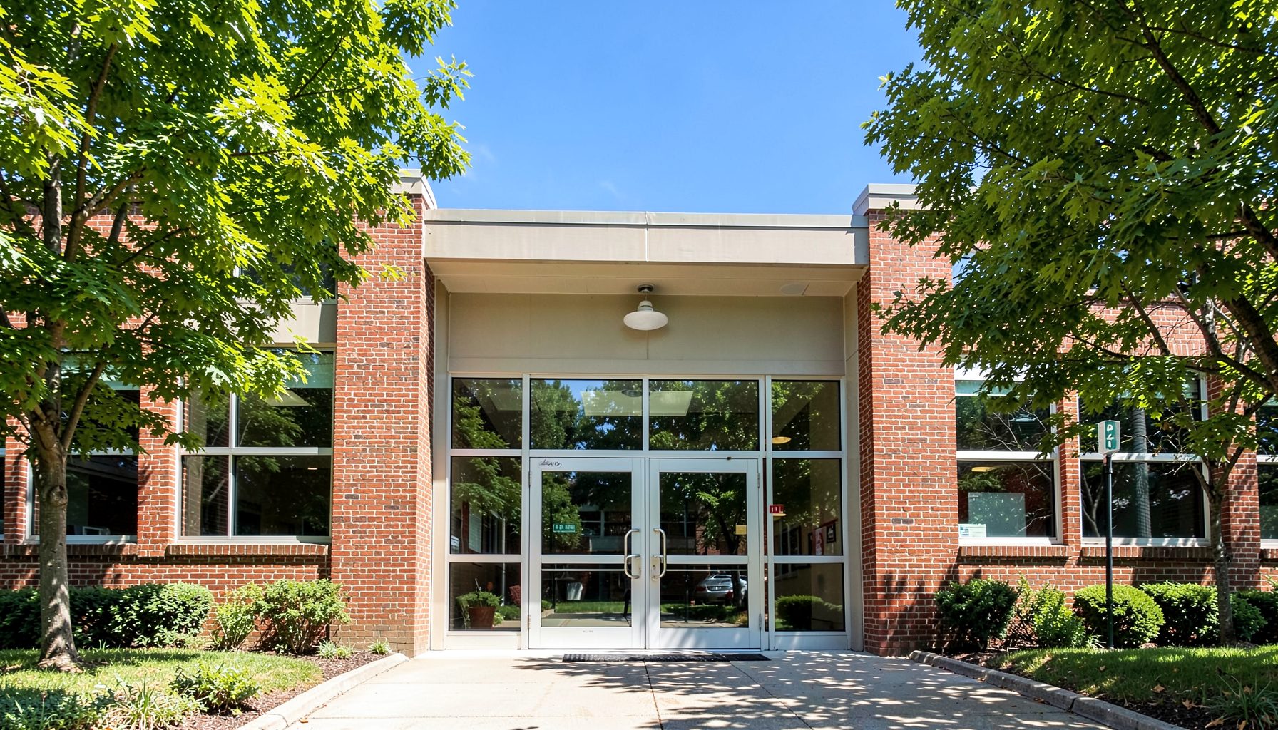 A sunlit brick school building entrance with wide glass doors, framed by green trees and a clear blue sky, conveying a welcoming and warm atmosphere