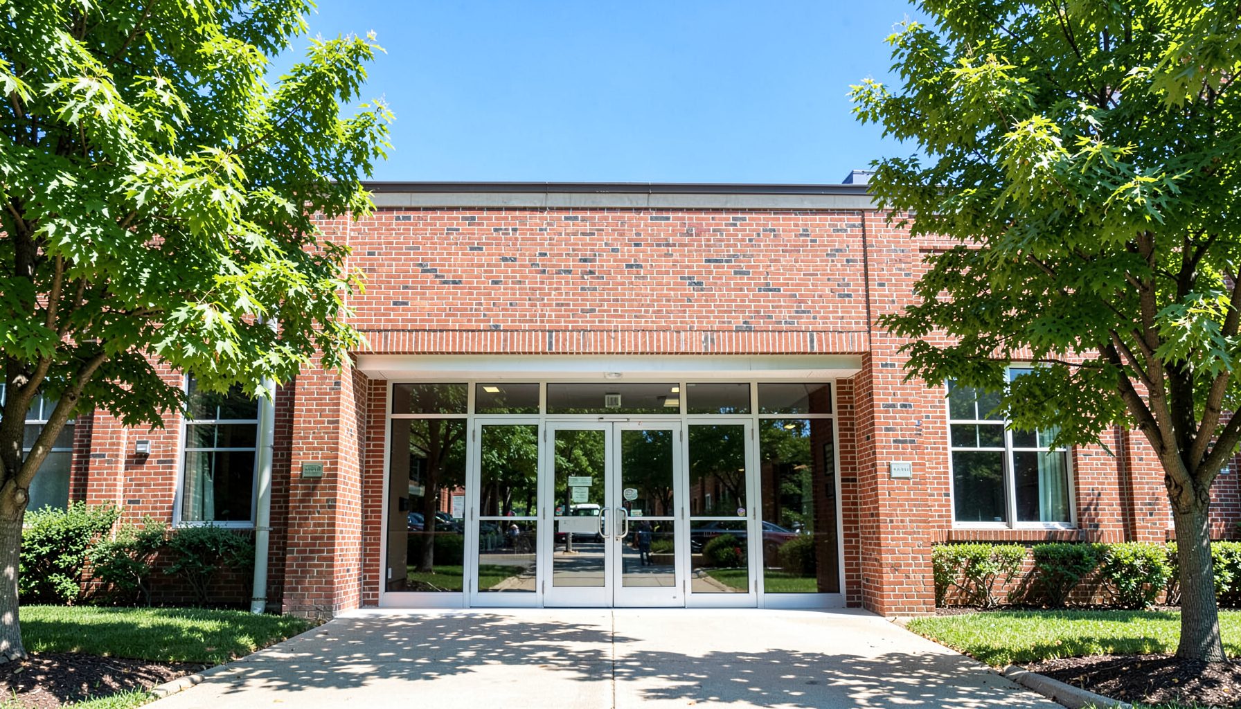 A sunlit brick school building entrance with wide glass doors, framed by green trees and a clear blue sky, conveying a welcoming and hopeful atmosphere